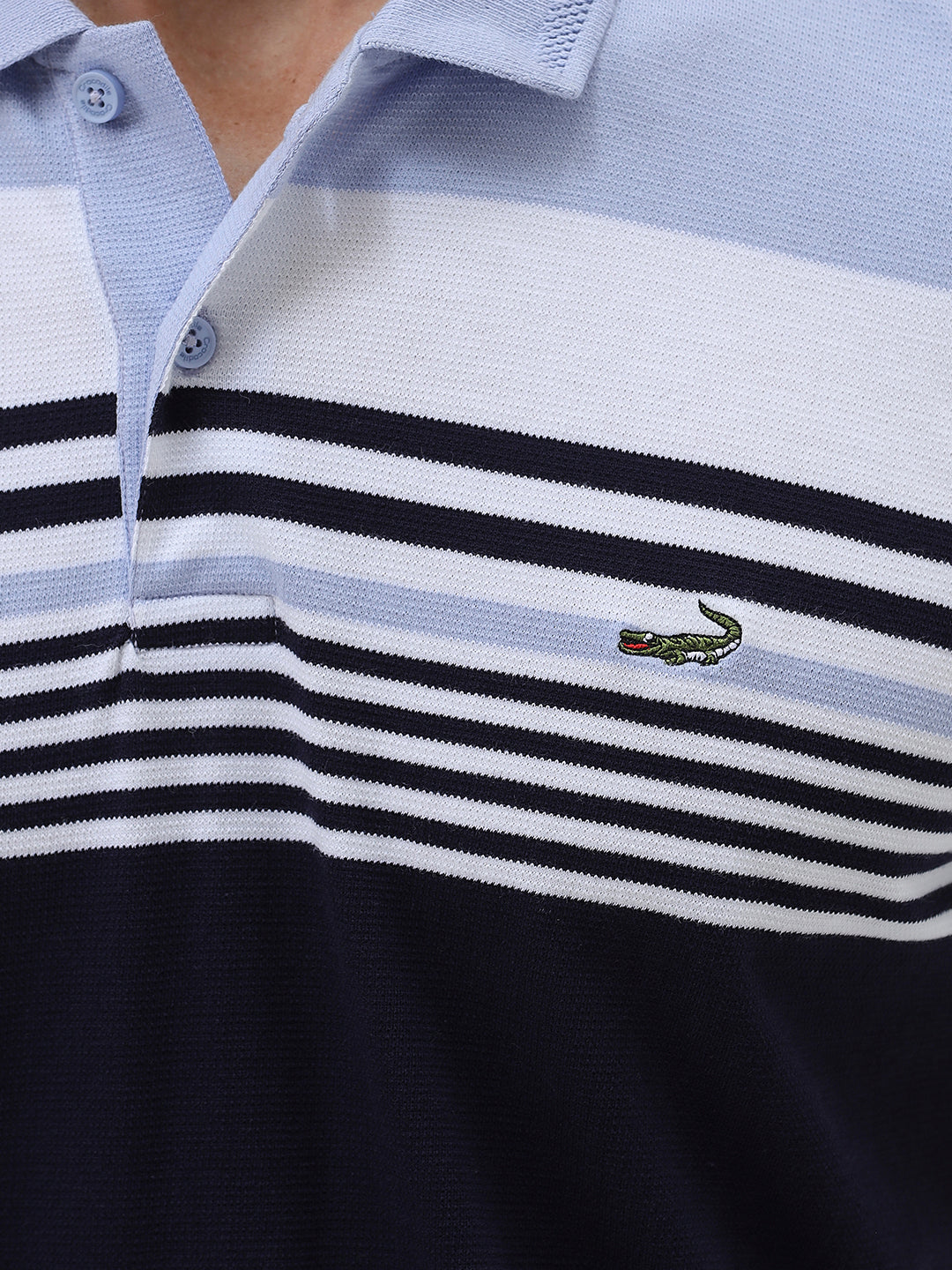 A man wearing a short-sleeve polo shirt with horizontal stripes in light blue, white, and navy, paired with light gray trousers, against a plain light gray background.