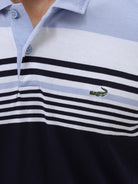 A man wearing a short-sleeve polo shirt with horizontal stripes in light blue, white, and navy, paired with light gray trousers, against a plain light gray background.