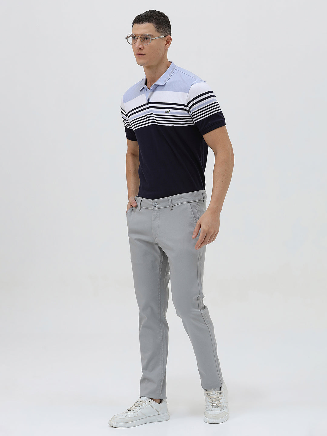 A man wearing a short-sleeve polo shirt with horizontal stripes in light blue, white, and navy, paired with light gray trousers, against a plain light gray background.