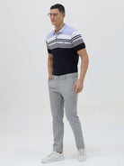 A man wearing a short-sleeve polo shirt with horizontal stripes in light blue, white, and navy, paired with light gray trousers, against a plain light gray background.