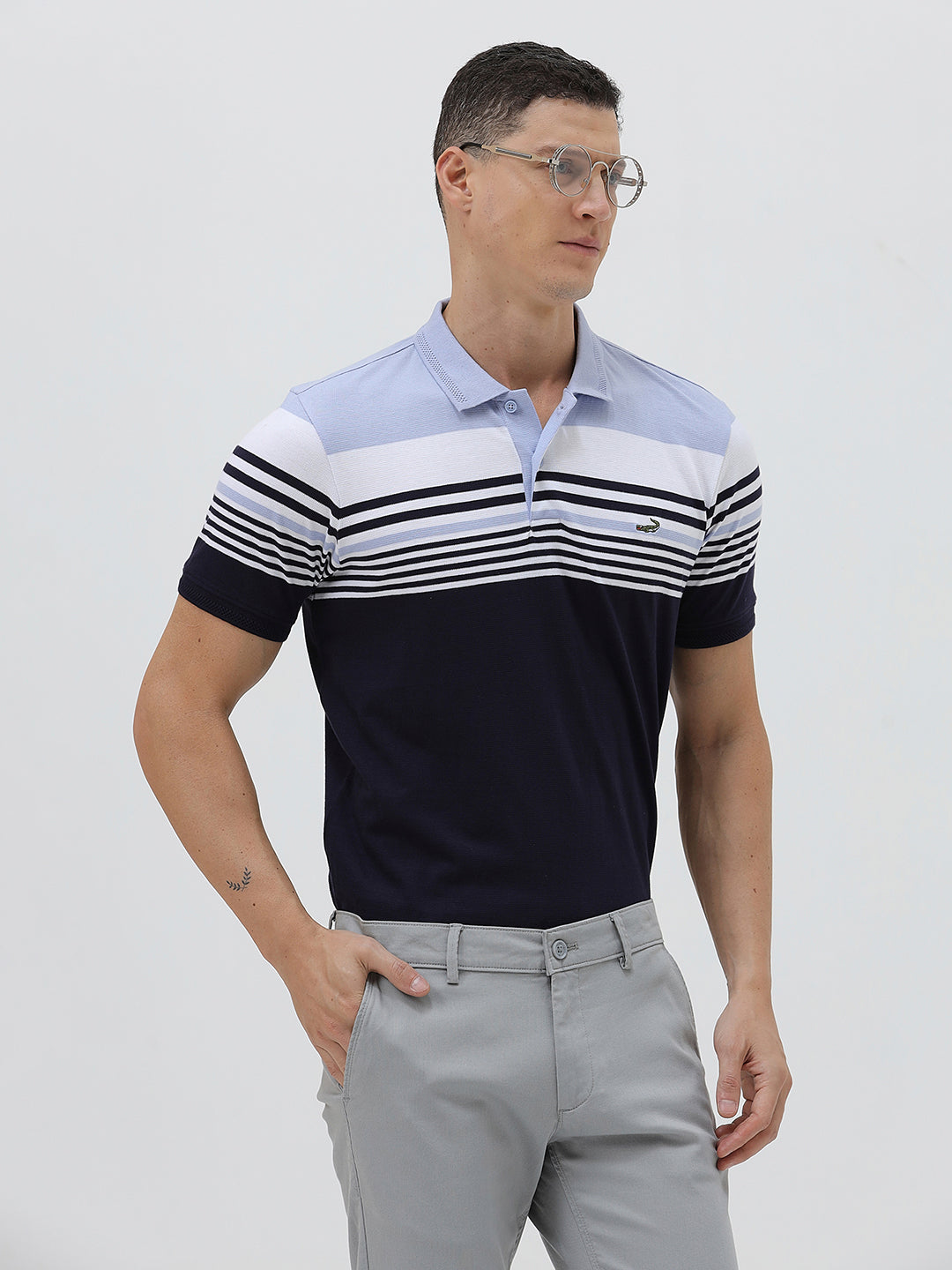 A man wearing a short-sleeve polo shirt with horizontal stripes in light blue, white, and navy, paired with light gray trousers, against a plain light gray background.