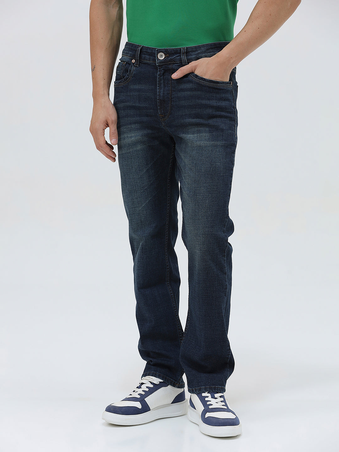 A person wearing dark blue slim-fit jeans, a green shirt, and navy and white sneakers, standing against a light gray studio background.