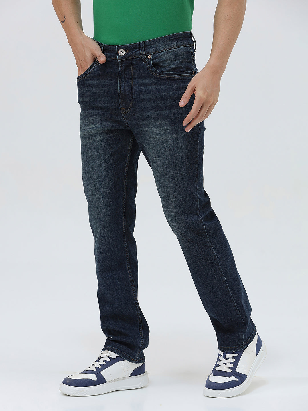 A person wearing dark blue slim-fit jeans, a green shirt, and navy and white sneakers, standing against a light gray studio background.