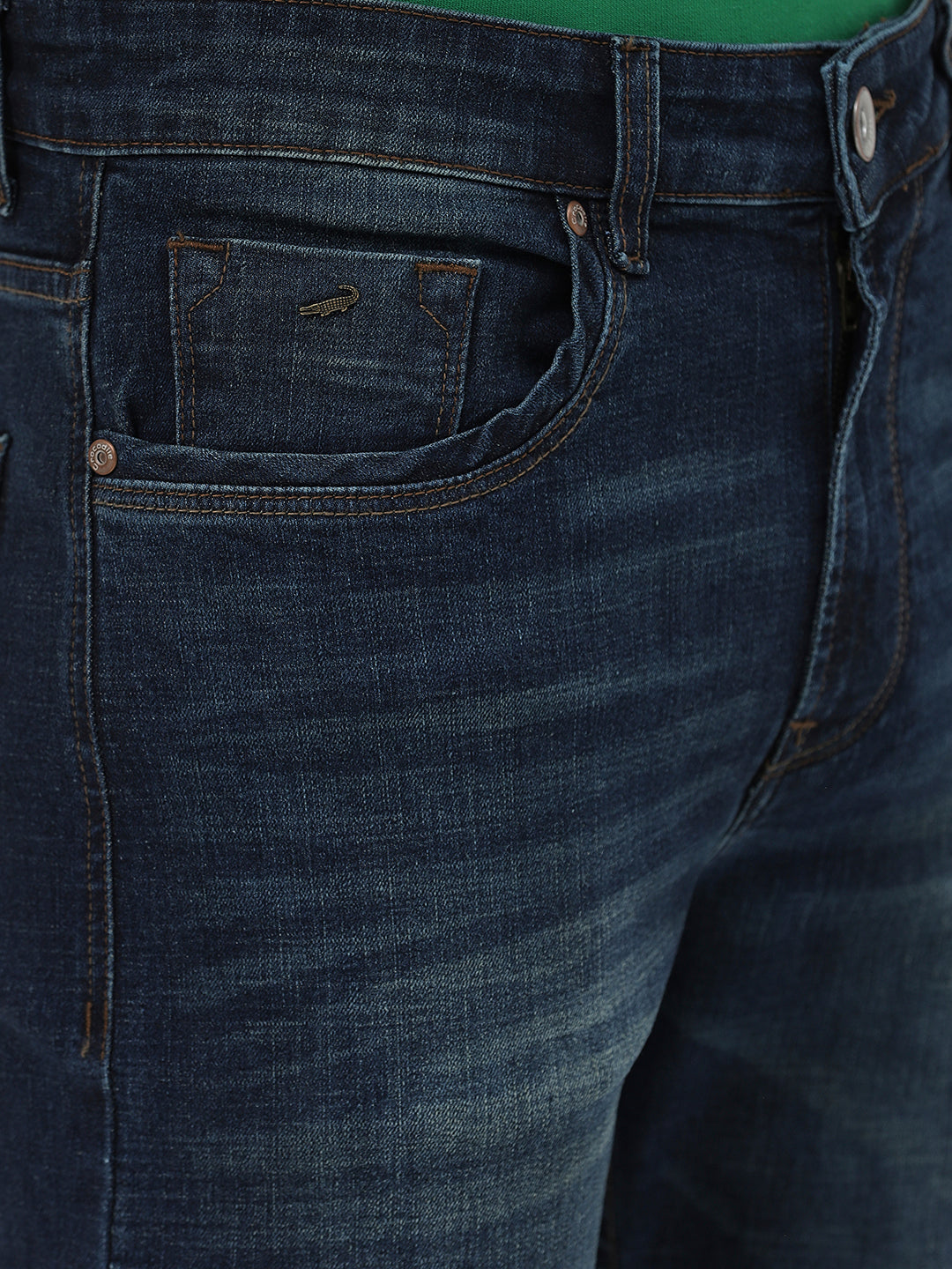 A person wearing dark blue slim-fit jeans, a green shirt, and navy and white sneakers, standing against a light gray studio background.