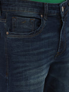A person wearing dark blue slim-fit jeans, a green shirt, and navy and white sneakers, standing against a light gray studio background.