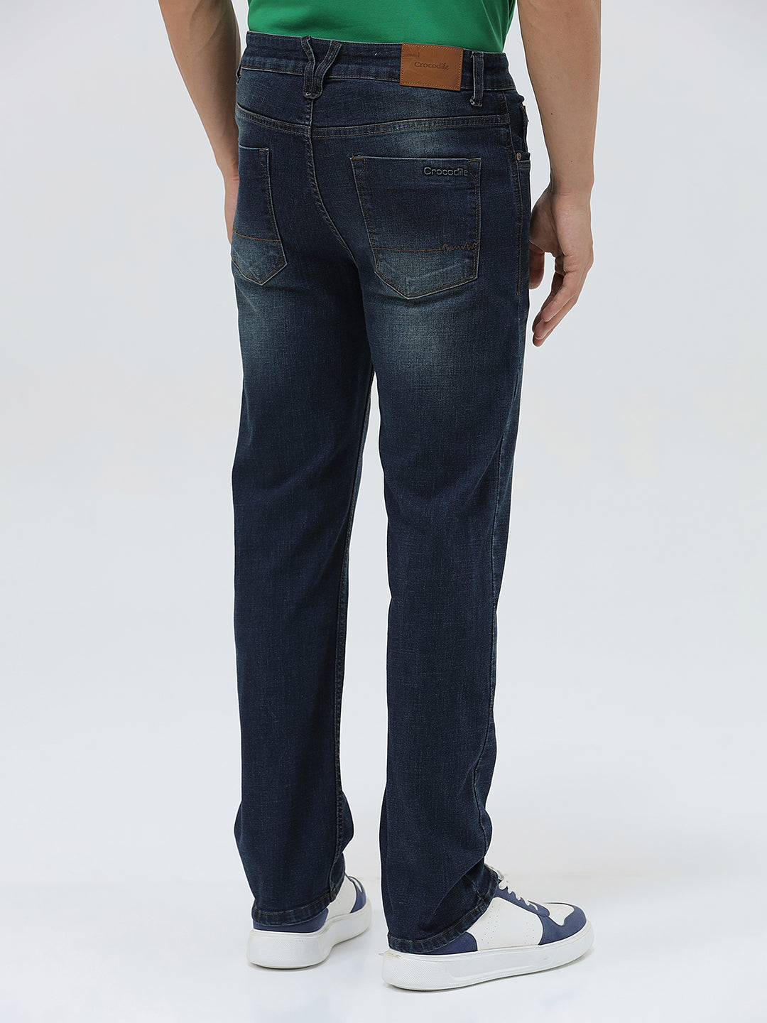 A person wearing dark blue slim-fit jeans, a green shirt, and navy and white sneakers, standing against a light gray studio background.