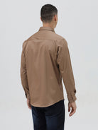 A man wearing a brown long-sleeve button-down shirt and dark blue jeans, standing against a light gray studio background.