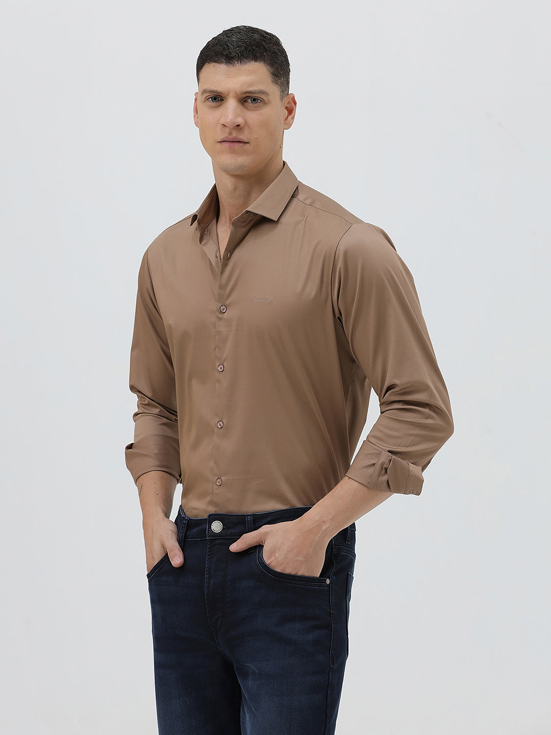 A man wearing a brown long-sleeve button-down shirt and dark blue jeans, standing against a light gray studio background.