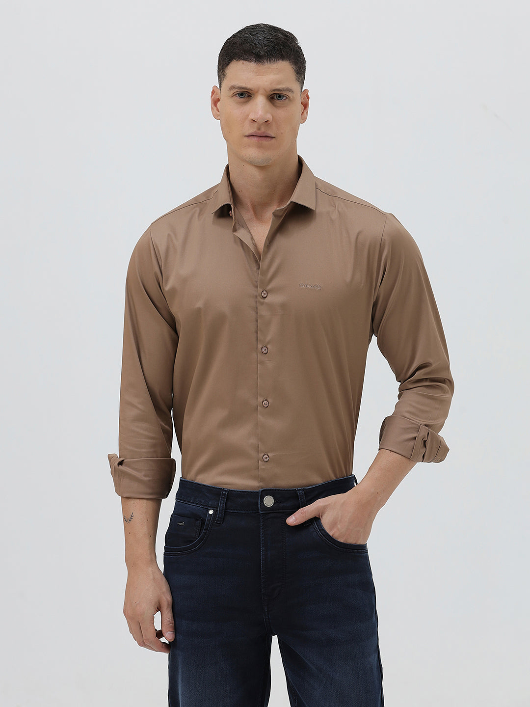 A man wearing a brown long-sleeve button-down shirt and dark blue jeans, standing against a light gray studio background.