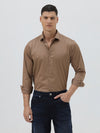 A man wearing a brown long-sleeve button-down shirt and dark blue jeans, standing against a light gray studio background.