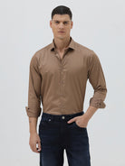 A man wearing a brown long-sleeve button-down shirt and dark blue jeans, standing against a light gray studio background.