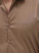 A man wearing a brown long-sleeve button-down shirt and dark blue jeans, standing against a light gray studio background.