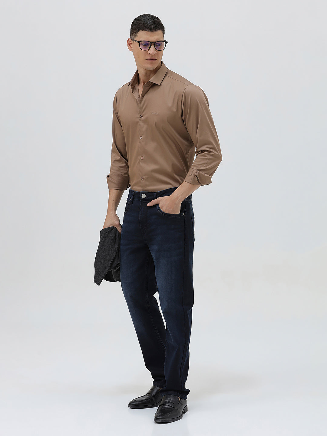 A man wearing a brown long-sleeve button-down shirt and dark blue jeans, standing against a light gray studio background.