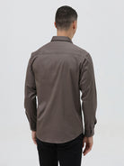 A man wearing a long-sleeve button-down dress shirt in a dark taupe-brown color, paired with black trousers, photographed against a light gray studio background.