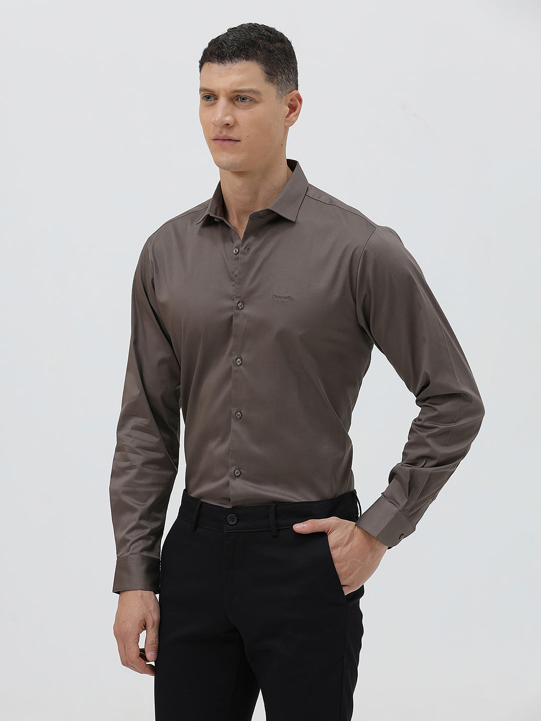A man wearing a long-sleeve button-down dress shirt in a dark taupe-brown color, paired with black trousers, photographed against a light gray studio background.