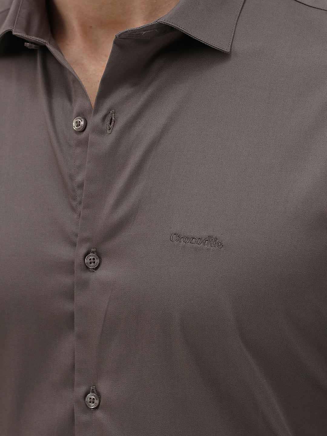 A man wearing a long-sleeve button-down dress shirt in a dark taupe-brown color, paired with black trousers, photographed against a light gray studio background.