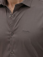 A man wearing a long-sleeve button-down dress shirt in a dark taupe-brown color, paired with black trousers, photographed against a light gray studio background.