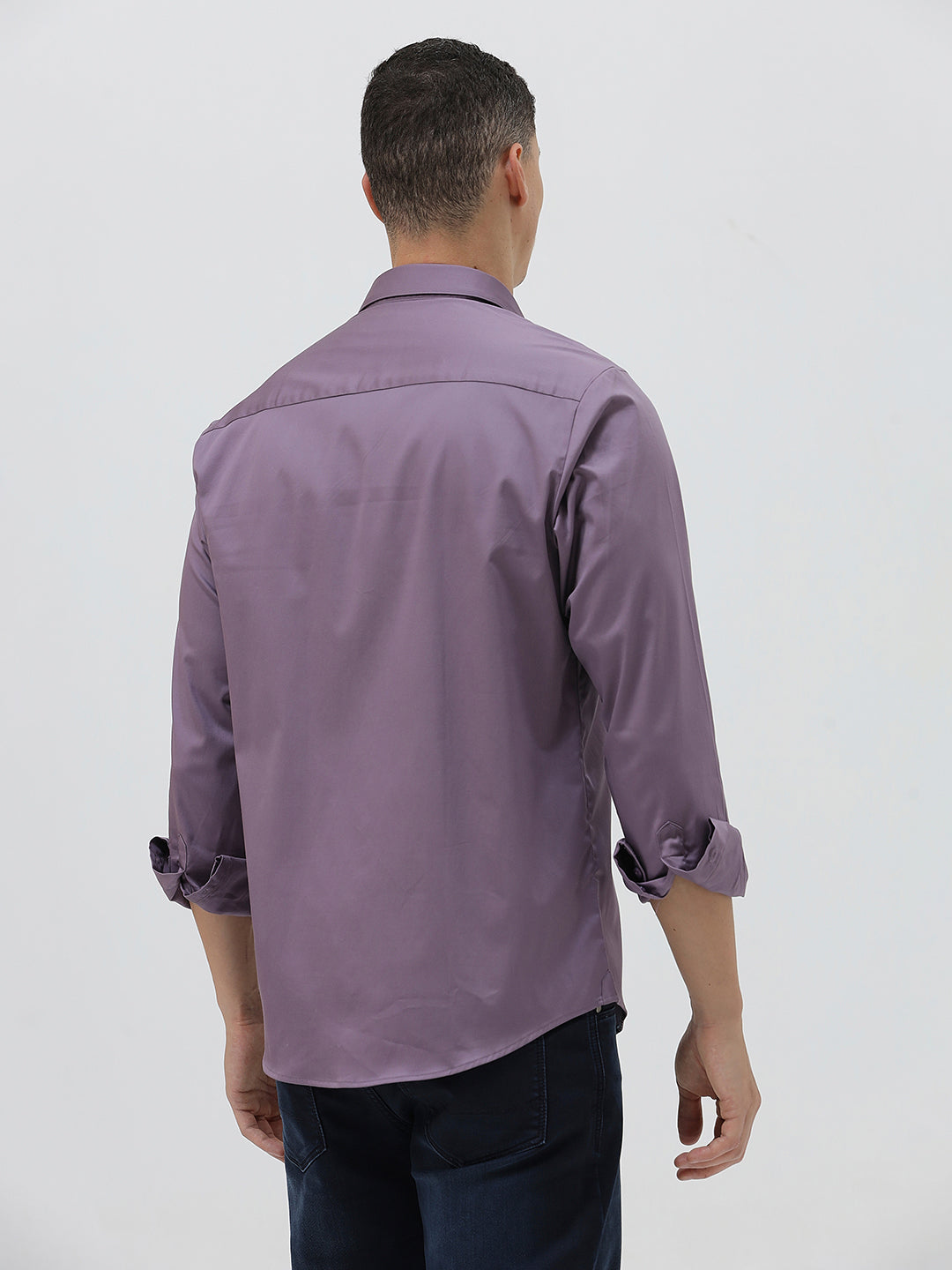 A man wearing a long-sleeve button-down shirt in a muted purple shade, paired with dark blue jeans, standing against a light gray studio background.