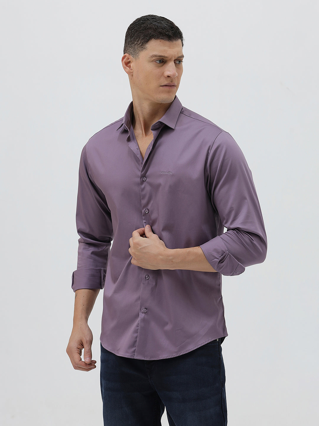 A man wearing a long-sleeve button-down shirt in a muted purple shade, paired with dark blue jeans, standing against a light gray studio background.