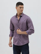 A man wearing a long-sleeve button-down shirt in a muted purple shade, paired with dark blue jeans, standing against a light gray studio background.