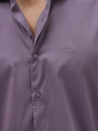 A man wearing a long-sleeve button-down shirt in a muted purple shade, paired with dark blue jeans, standing against a light gray studio background.