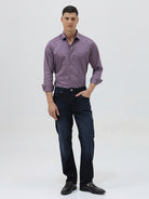 A man wearing a long-sleeve button-down shirt in a muted purple shade, paired with dark blue jeans, standing against a light gray studio background.