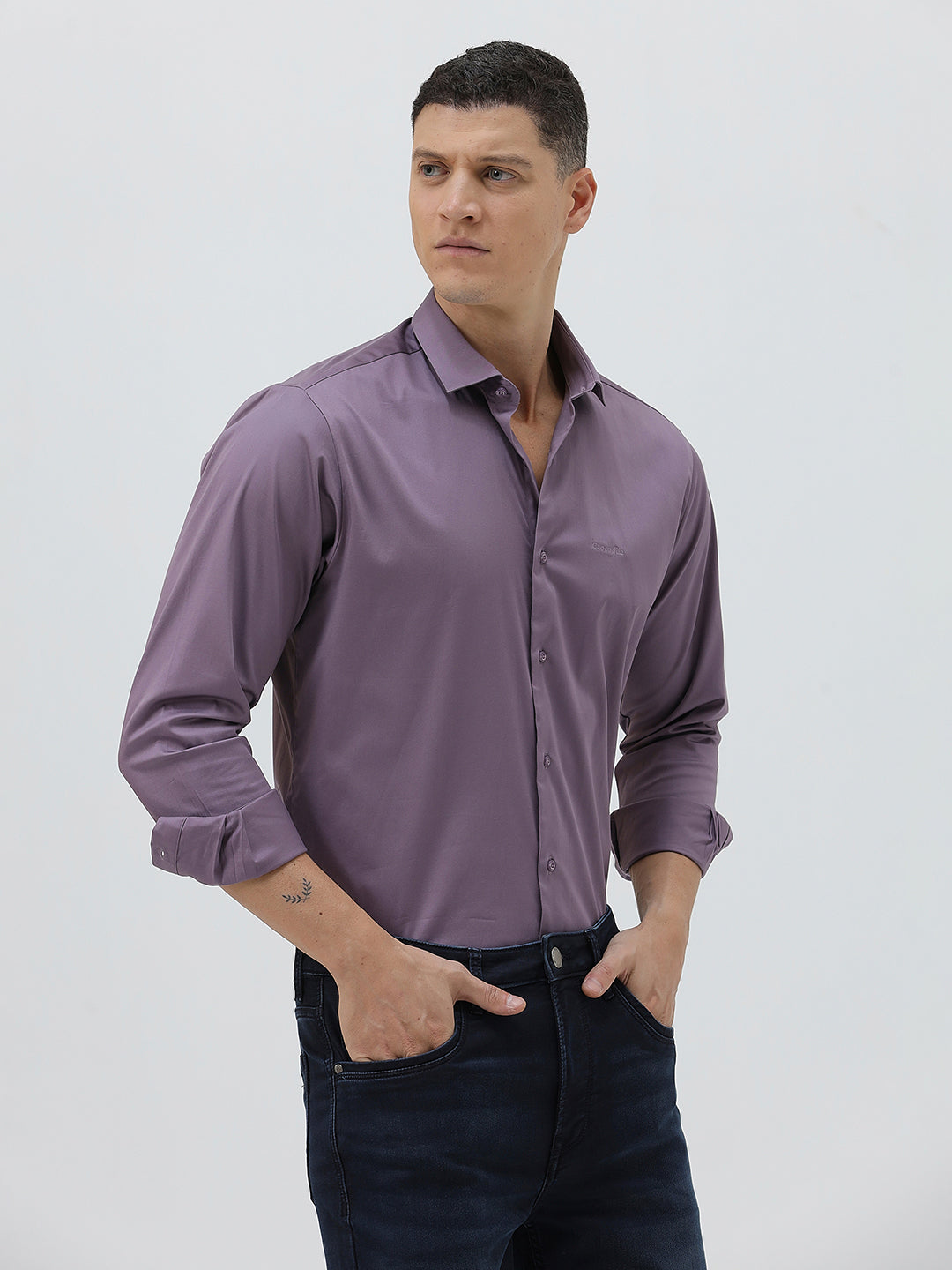 A man wearing a long-sleeve button-down shirt in a muted purple shade, paired with dark blue jeans, standing against a light gray studio background.