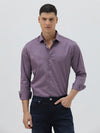 A man wearing a long-sleeve button-down shirt in a muted purple shade, paired with dark blue jeans, standing against a light gray studio background.