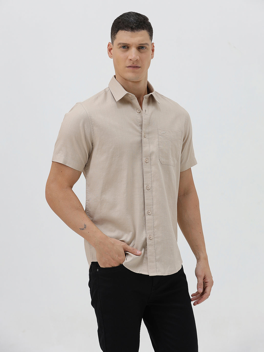 A man wearing a dusty rose long-sleeve button-down shirt and light beige trousers, standing against a light gray studio background.