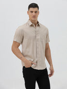 A man wearing a dusty rose long-sleeve button-down shirt and light beige trousers, standing against a light gray studio background.
