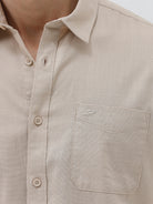 A man wearing a dusty rose long-sleeve button-down shirt and light beige trousers, standing against a light gray studio background.