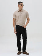 A man wearing a dusty rose long-sleeve button-down shirt and light beige trousers, standing against a light gray studio background.