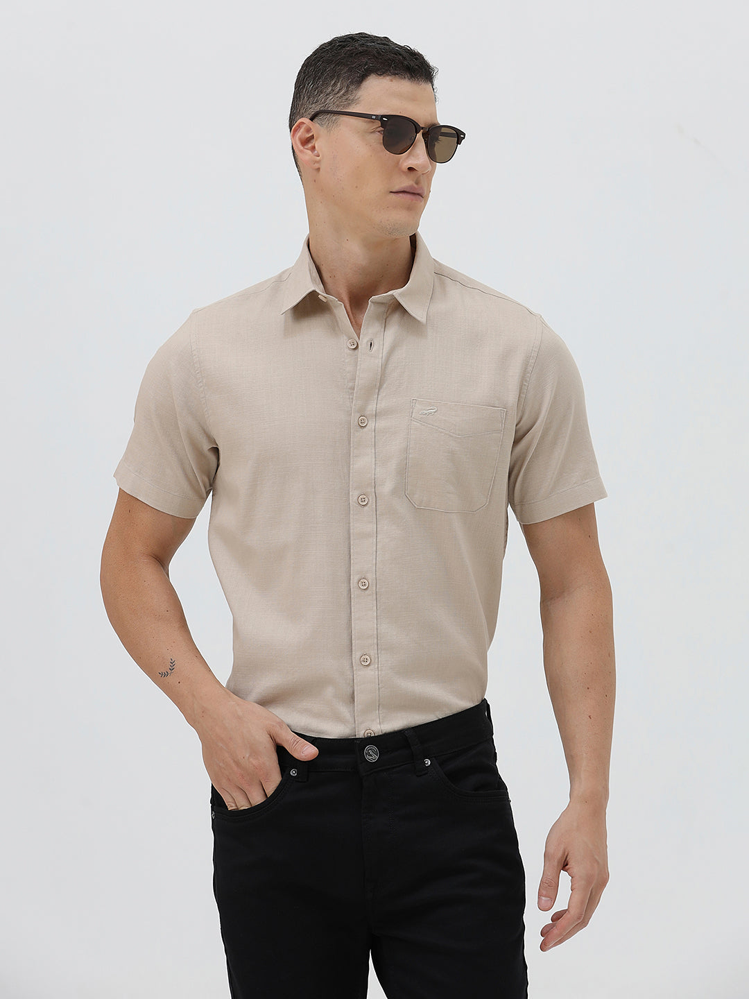 A man wearing a dusty rose long-sleeve button-down shirt and light beige trousers, standing against a light gray studio background.