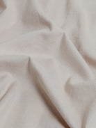 A man wearing a dusty rose long-sleeve button-down shirt and light beige trousers, standing against a light gray studio background.