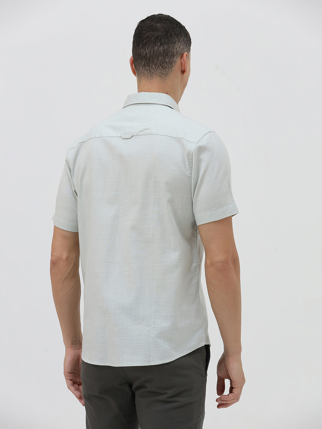A man wearing a light sage green short-sleeve button-down shirt and dark gray trousers, standing against a plain light gray studio background.