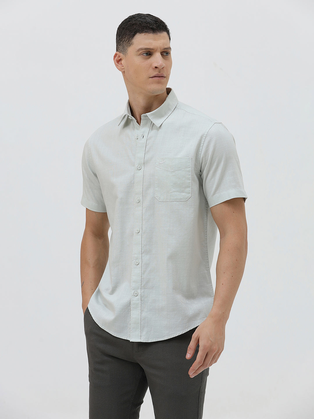 A man wearing a light sage green short-sleeve button-down shirt and dark gray trousers, standing against a plain light gray studio background.