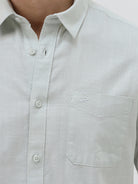 A man wearing a light sage green short-sleeve button-down shirt and dark gray trousers, standing against a plain light gray studio background.