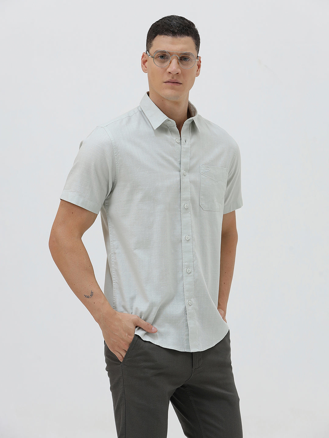 A man wearing a light sage green short-sleeve button-down shirt and dark gray trousers, standing against a plain light gray studio background.