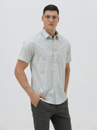 A man wearing a light sage green short-sleeve button-down shirt and dark gray trousers, standing against a plain light gray studio background.