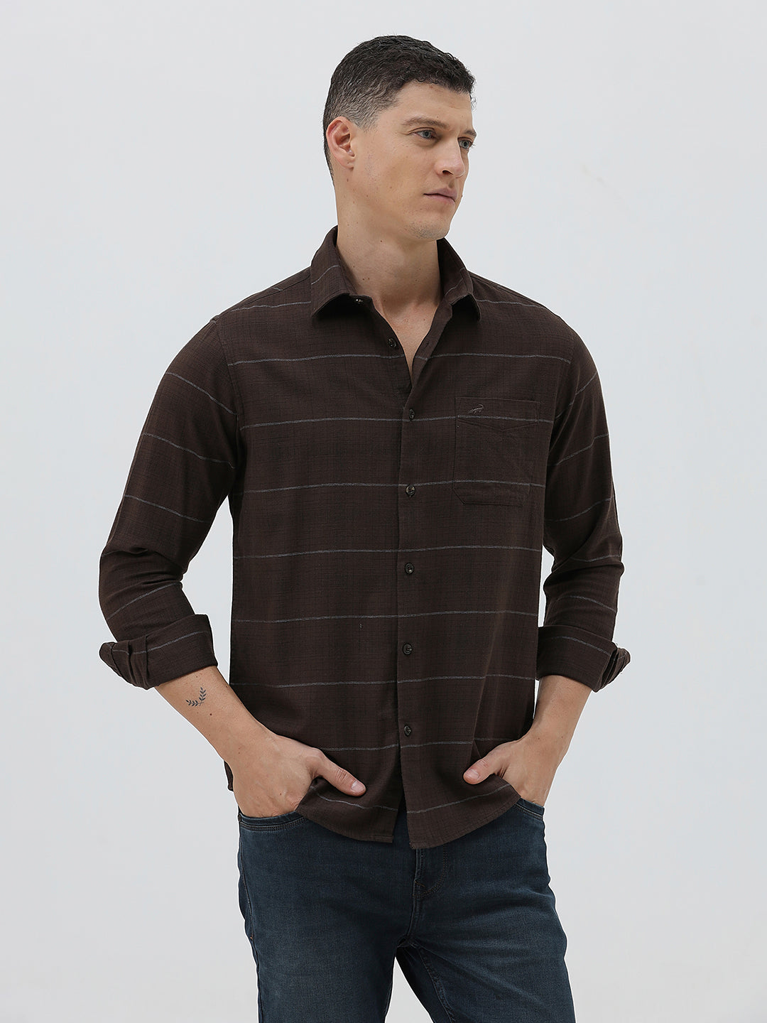 A man wearing a dark brown long-sleeve button-up shirt with thin horizontal stripes, paired with dark blue jeans, standing against a light gray studio background.