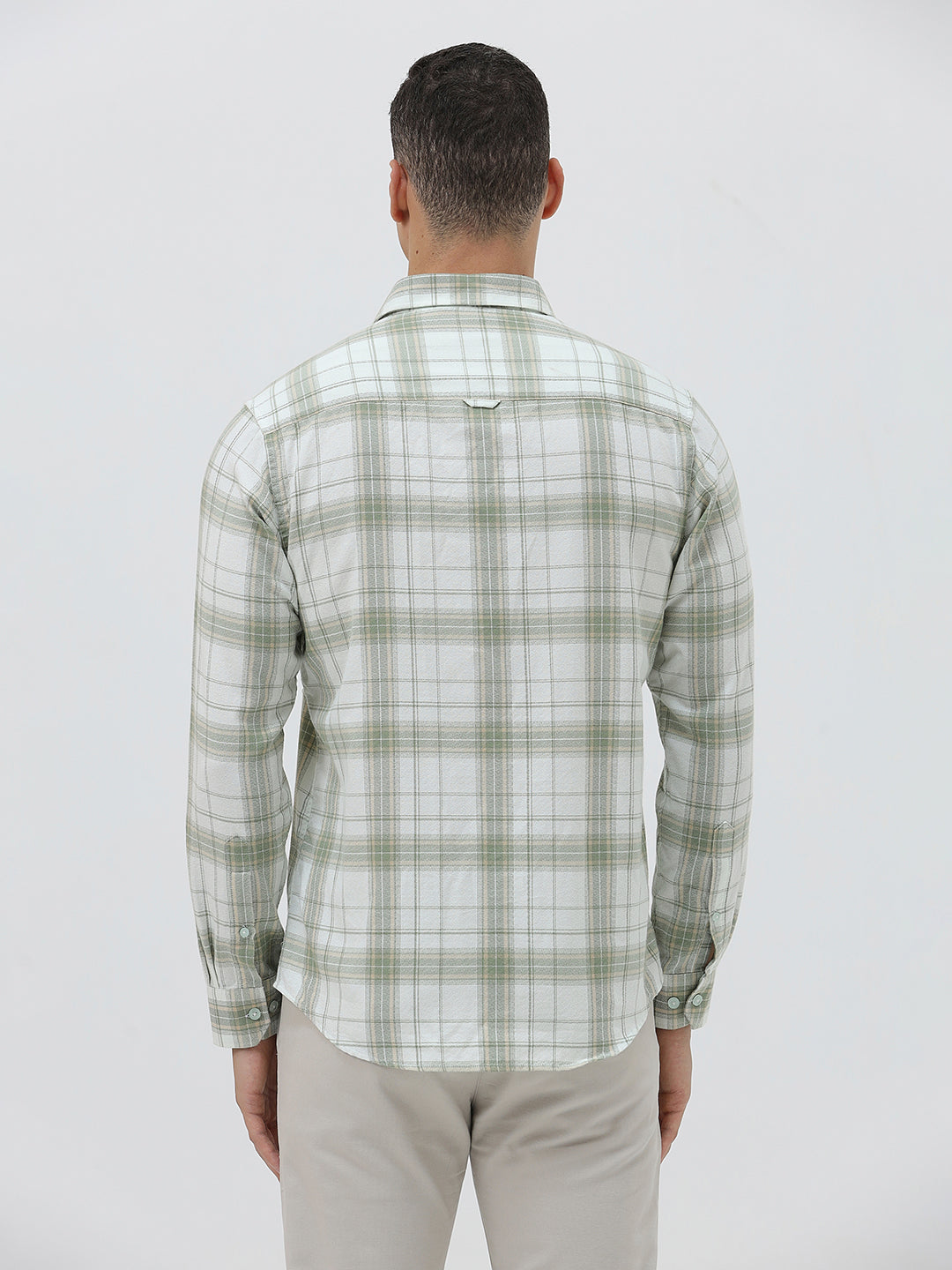 A man wearing a light green and white plaid long-sleeve button-up shirt with beige trousers, standing against a plain light gray background.