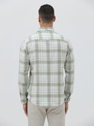 A man wearing a light green and white plaid long-sleeve button-up shirt with beige trousers, standing against a plain light gray background.