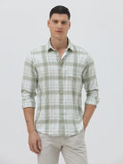 A man wearing a light green and white plaid long-sleeve button-up shirt with beige trousers, standing against a plain light gray background.