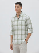 A man wearing a light green and white plaid long-sleeve button-up shirt with beige trousers, standing against a plain light gray background.