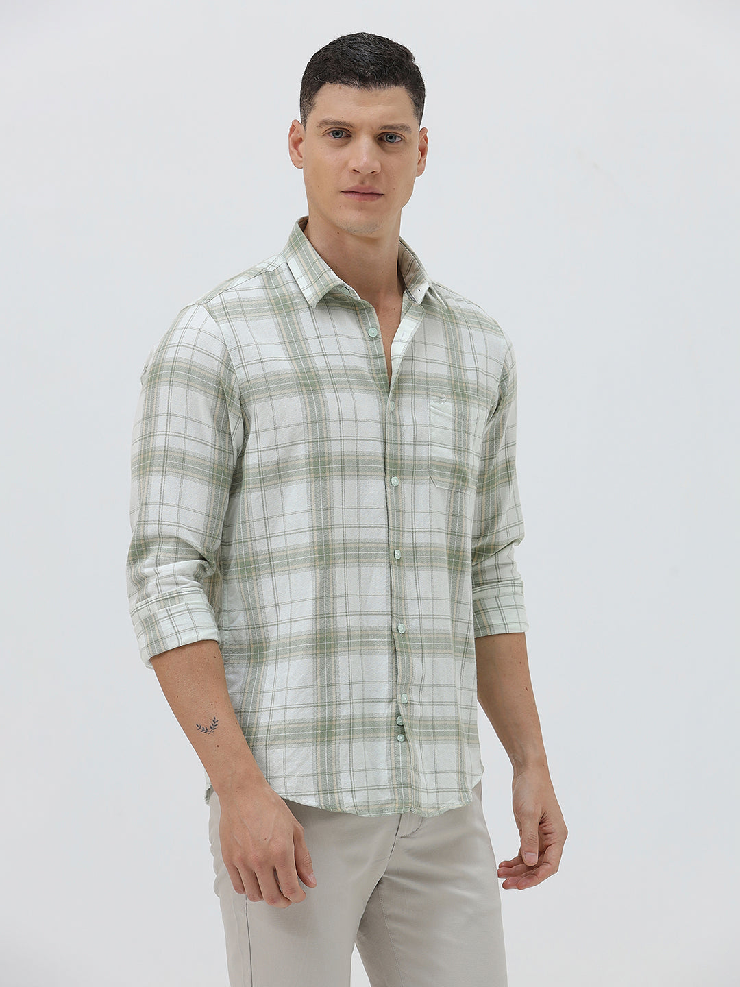 A man wearing a light green and white plaid long-sleeve button-up shirt with beige trousers, standing against a plain light gray background.