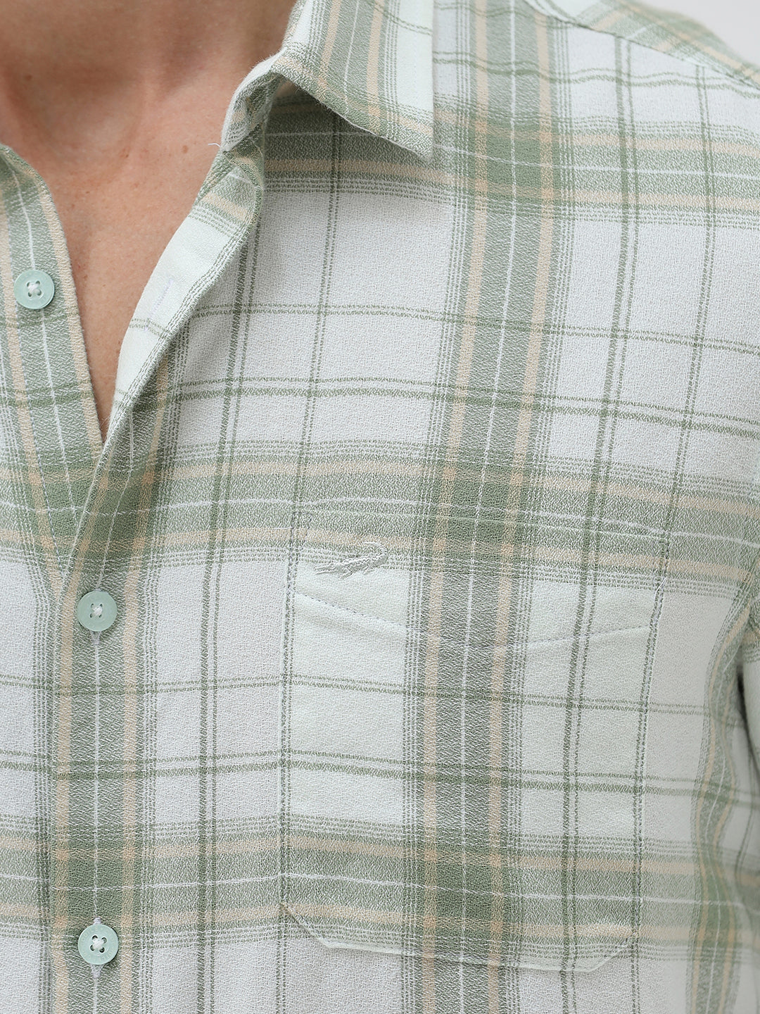 A man wearing a light green and white plaid long-sleeve button-up shirt with beige trousers, standing against a plain light gray background.