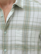 A man wearing a light green and white plaid long-sleeve button-up shirt with beige trousers, standing against a plain light gray background.