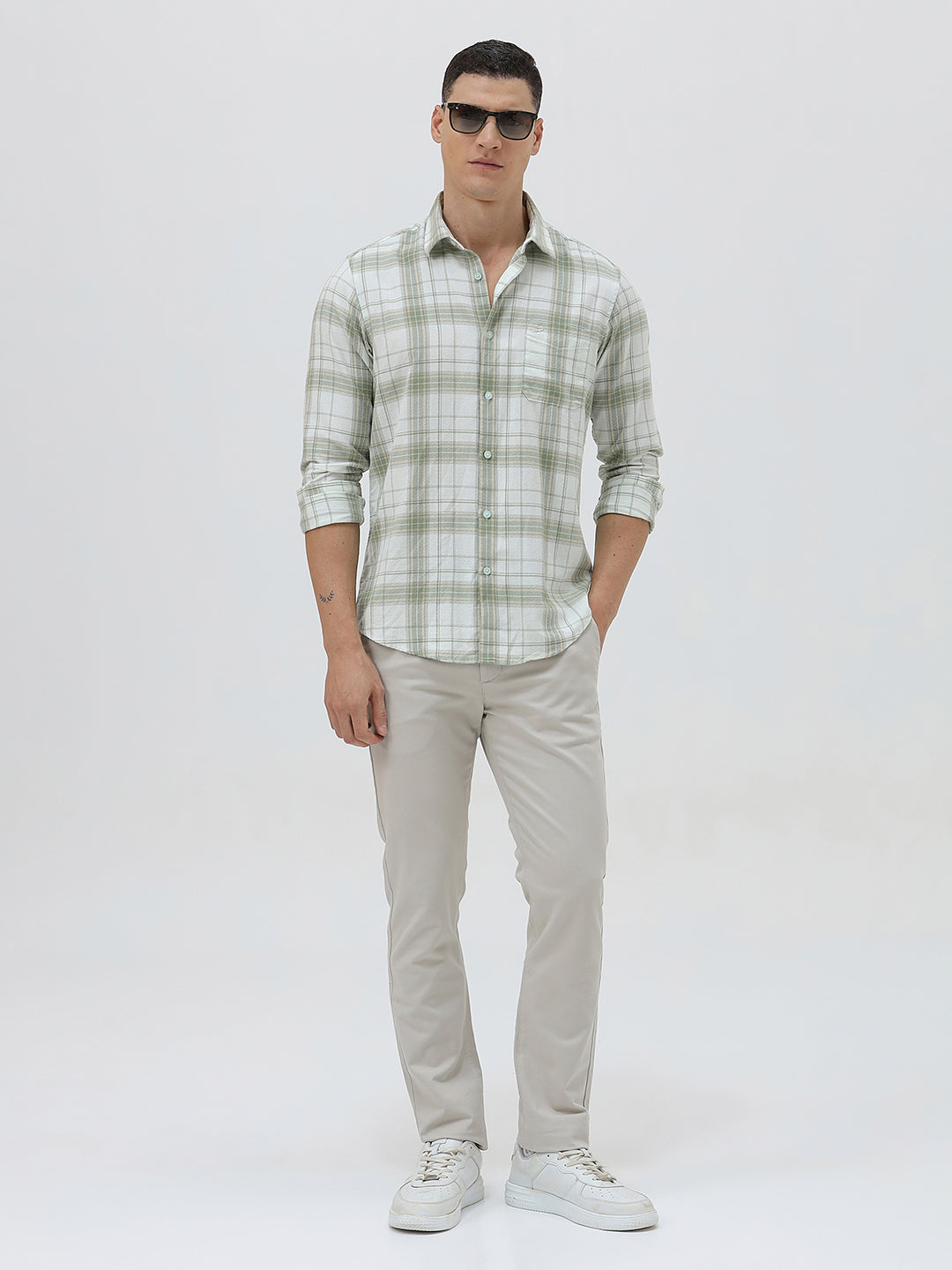 A man wearing a light green and white plaid long-sleeve button-up shirt with beige trousers, standing against a plain light gray background.