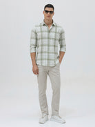A man wearing a light green and white plaid long-sleeve button-up shirt with beige trousers, standing against a plain light gray background.