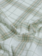 A man wearing a light green and white plaid long-sleeve button-up shirt with beige trousers, standing against a plain light gray background.